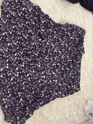 Lila blommig kortkjol - Söt kortkjol med blommigt mönster i lila, vitt och svart. Kjolen har en lätt volangig kant och är gjord i ett tunt, luftigt material som passar perfekt till varmare dagar. Elastisk midja för skön passform.