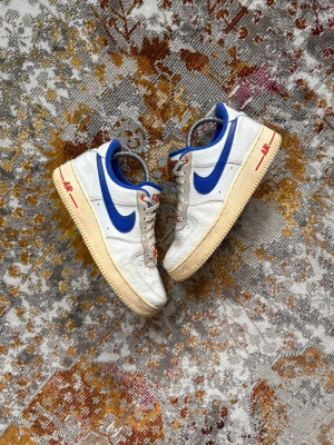 Nike Air Force 1 vita/blå sneakers - Nike Air Force 1 sneakers i vitt läder med blå swoosh och detaljer. Yttersulan är beige och har röda 'AIR'-detaljer på sidan. Skon har blå vadderad häl och klassisk låg siluett. Perfekt för dig som gillar retrovibbar och streetstyle.