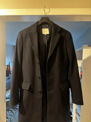 Svart ullkappa från Studio Total - Stilren svart kappa från Studio Total i klassisk rak modell. Kappan har tre knappar framtill, två fickor med lock och slät finish. Storlek 46.