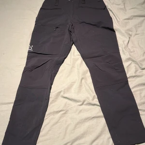 Svarta friluftsbyxor från Haglöfs - Svarta friluftsbyxor från Haglöfs med flera fickor och dragkedja på ena benet. Byxorna har en rak passform och är tillverkade i slitstarkt syntetmaterial, perfekta för utomhusaktiviteter.