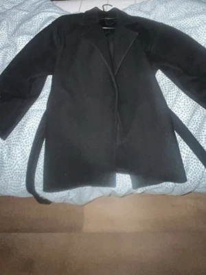 Svart klassisk kappa med skärp från Missäy  - Stilren svart kappa med klassisk krage och tillhörande midjebälte. Kappan har rak passform och långa ärmar, perfekt för kyligare dagar. Materialet ser ut att vara ullblandning för extra värme och komfort. Enkel och tidlös design som passar till det mesta.