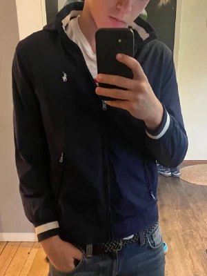 Mörkblå vindjacka från Ralph Lauren - Snygg mörkblå vindjacka från Ralph Lauren med dragkedja framtill och vita detaljer vid ärmslut och krage. Jackan har två fickor med dragkedja och den klassiska loggan på bröstet. Perfekt för en stilren och sportig look. Storleken är L ungdom motsvarar Xs