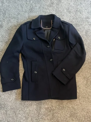 Marinblå rock med fickdetaljer - Stilren marinblå rock med klassisk krage, fyra fickor framtill och knappdetaljer på ärmar och axlar. Rocken har en rak passform och är fodrad för extra komfort. Perfekt för dig som vill ha en snygg och tidlös look.