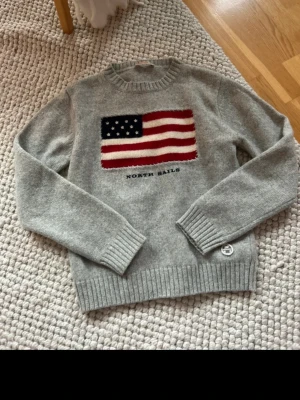 Grå stickad tröja North Sails - Snygg grå stickad tröja från North Sails med amerikansk flagga på bröstet. Tröjan har rund hals, ribbstickade muddar och nederkant samt en liten patch vid midjan. Perfekt för dig som gillar klassisk och sportig stil. Står inte stolek men antar s 