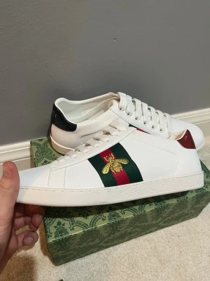 Gucci Ace Bee vita sneakers - Säljer ett par vita Gucci Ace sneakers med klassiska gröna och röda ränder samt broderad guldbi på sidan. Skorna har läderdetaljer i rött och grönt med ormmönster på hälen och vita snören. Perfekta för dig som vill ha en lyxig och stilren look.helt nya köpa på Plick men var tyvärr för små vet ej om dom e äkta 