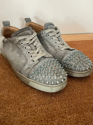 Gråa Louboutin skor - Unika gråa sneakers i mocka med coola silverfärgade nitar över tån. Skorna har vita snören och en klassisk låg siluett. Insidan är beige och sulan är vit. Perfekta för dig som vill sticka ut med din stil. (Dem har tappat en spik och har varit hos skomakaren en gång)