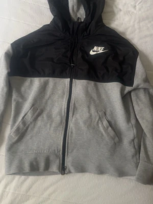 Nike hoodie med dragkedja, grå/svart - Nike hoodie i storlek S med dragkedja framtill. Färgen är grå med svart upptill och vit Nike-logga på bröstet. Två fickor på sidorna och justerbar huva. Tillverkad i mjukt material som känns skönt mot huden. Perfekt för en sportig och avslappnad stil.