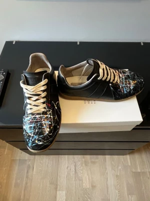 Svarta sneakers från Maison Margiela - Unika svarta sneakers från Maison Margiela med färgstänk i blått, gult, vitt och rött över tå och sidor. Skorna har beige sula och vita snören. Tillverkade i läder med klassisk låg siluett och diskreta paneldetaljer. Perfekt för dig som gillar statement-plagg.