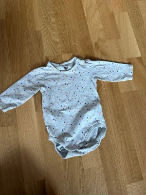 Grå body med färgglada droppar Åhléns - Supersöt grå långärmad body från Åhléns med små färgglada droppar i rosa, blått, gult och orange. Praktiska tryckknappar vid axeln och i grenen gör den enkel att ta på och av. Perfekt för mysiga dagar hemma.