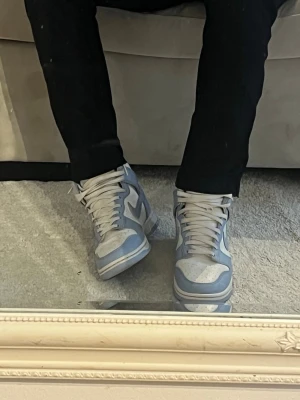 Nike Dunk High blå och vita sneakers - Nike Dunk High sneakers i ljusblått och vitt läder med klassisk Swoosh på sidan. Högt skaft, vita skosnören och perforerad tåbox för extra ventilation. Snygg retrostil som passar perfekt till streetwear och casual outfits.