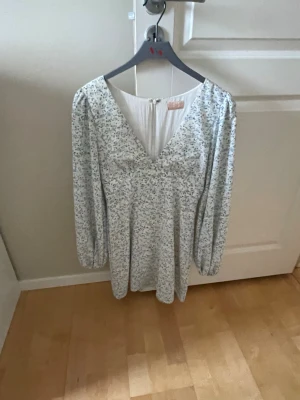 Vit blommig kortklänning med v-ringning - Söt vit kortklänning med små blommor, v-ringad framtill och långa ärmar. Klänningen har en lätt utställd passform och diskret dragkedja i ryggen. Perfekt för sommarens alla tillfällen.