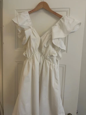 Vit kortklänning med volang och rosett - Supersöt vit kortklänning med volangdetaljer vid axlarna och stor rosett i ryggen. Klänningen har v-ringning och är ärmlös, 💛 