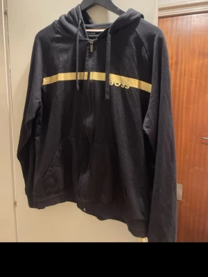 Svart hoodie från BOSS - Snygg svart hoodie från BOSS med dragkedja framtill och en guldfärgad rand över bröstet samt BOSS-logga. Hoodien har huva med dragsko och två fickor framtill. Perfekt för en chill och stilren look.