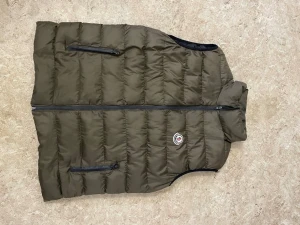 Olivgrön dunväst från Moncler - Snygg olivgrön dunväst från Moncler med två dragkedjefickor framtill och klassisk Moncler-logga på bröstet. Västen har hög krage och quiltad design som ger en sportig och trendig look. Perfekt att slänga över en hoodie eller tröja för extra värme.