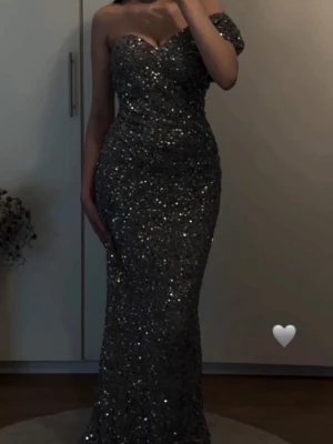 Glittrig silver långklänning oneshoulder - Supersnygg långklänning i silver med massor av glitter. Klänningen har en figurnära passform och en axelbandslös design med en axel täckt, vilket ger en elegant oneshoulder-look. Perfekt för bal eller festliga tillfällen.