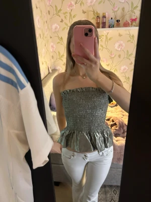 Silvrig/flerfärgad bandeau tubtopp med volang - Säljer en silvrig bandeau tubtopp med smockad överdel och volang nertill. Toppen är axelbandslös och har en glansig finish som ger en riktigt snygg look. Perfekt för dig som vill sticka ut lite extra.