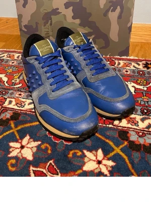 Valentino Rockrunner 🎸🤘 - Snygga blå sneakers från Valentino med grå detaljer och blå skosnören. Skorna har coola nitar på sidan och en beige sula. Ovandelen är i en mix av läder och mocka, och insidan är svart. Perfekta för dig som vill sticka ut med stilrena och exklusiva sneakers. (Det är en liten skada på hälen av ena skon.)👍