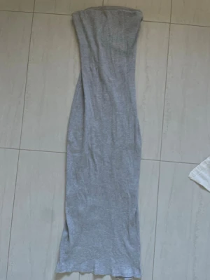 Grå axelbandslös långklänning - Snygg grå axelbandslös långklänning i ribbat material. Klänningen har en tight passform och en liten slits framtill för extra stilpoäng. Perfekt för dig som gillar minimalistisk och clean look.