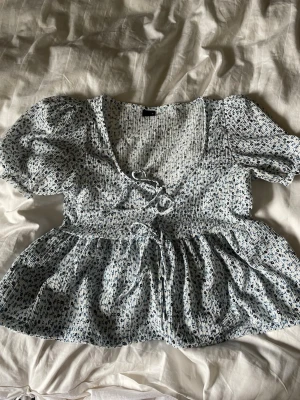 Blommig blus med puffärm och snörning från Gina tricot - Supersöt blommig blus med puffärmar och smockad överdel. Blusen har v-ringning, snörning framtill och volang nedtill. Tunt, luftigt material i vitt med blått blommönster – perfekt för vår och sommar.