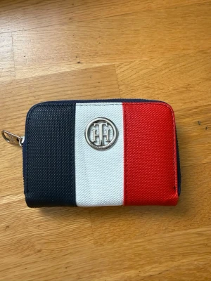 Randig plånbok från Tommy Hilfiger - Snygg plånbok från Tommy Hilfiger i textil med klassiska färgblock i marinblått, vitt och rött. Plånboken har ett silverfärgat TH-emblem på framsidan och dragkedja runt om. Perfekt storlek för kort och kontanter.