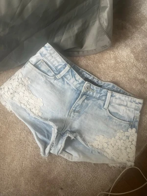 Ljusblå jeansshorts med spetsdetaljer - Säljer ett par ljusblå jeansshorts med vita spetsblommor på sidorna. Shortsen har fransig kant, fem fickor och bälteshällor. Perfekta för sommaren och ger en söt och avslappnad look.