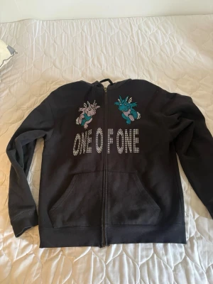 Svart hoodie med drakar och strass - Säljer en svart hoodie med dragkedja och huva. På bröstet står det 'ONE OF ONE' i strass och ovanför finns två färgglada drakar i blått och rosa, också i strass. Hoodien har känguruficka framtill och en avslappnad passform. Perfekt för dig som vill sticka ut.