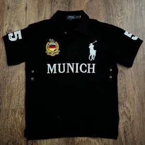 Chief keef Polo Ralph Lauren Munich t-shirt - Snygg svart t-shirt från Polo Ralph Lauren med stort vitt polospelar-tryck och texten 'MUNICH' på bröstet. T-shirten har korta ärmar, broderad logga och nummerdetaljer på ärmarna. Perfekt för dig som gillar sportig och klassisk stil.