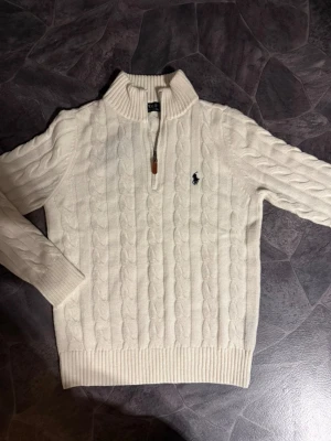 Ralph Lauren - Kabelstickad vit tröja från Polo Ralph Lauren med halv dragkedja och hög krage. Tröjan har det klassiska blå broderade logotypen på bröstet och ribbade muddar vid ärmslut och nederkant. Perfekt för lager på lager och snygg till jeans.