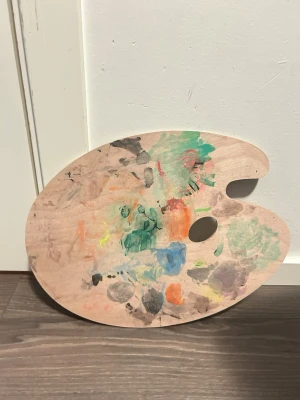 Oval träpalett för måleri - Säljer en klassisk oval konstnärspalett i trä med tumhål. Ena sidan har färgrester i grönt, blått, orange och svart medan andra sidan är helt ren. Perfekt för dig som gillar att måla och vill ha en smidig palett att blanda färger på.