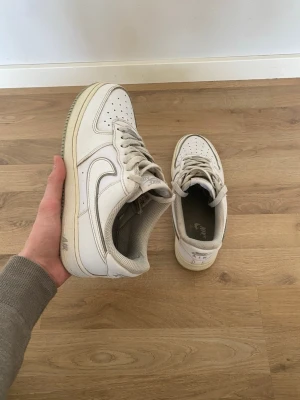 Vita Nike Air Force 1 sneakers - Klassiska vita Nike Air Force 1 sneakers med lågt skaft och ikonisk swoosh på sidan. Skorna har perforerad tå för ventilation, tjock sula och är tillverkade i läder med textilfoder. Perfekta för en clean och tidlös streetstil.