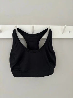 Svart sport-bh med brottarrygg - En stilren svart sport-bh med brottarrygg och meshdetaljer baktill för extra ventilation. Bh:n har bred resår under bysten och är tillverkad i ett stretchigt material som ger bra stöd vid träning. Perfekt för dig som vill ha en enkel och funktionell sport-bh.