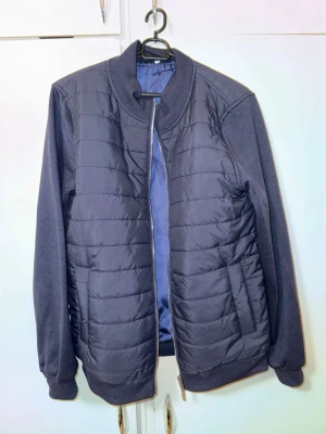 Mörkblå pufferjacka med dragkedja - Snygg mörkblå cardigan med quiltad framsida och släta ärmar. Jackan har ribbade muddar vid ärmslut och nederkant samt en hög krage. Stängs med dragkedja och har en lätt glansig insida. Perfekt för kalla dagar. Storlek M men passar S