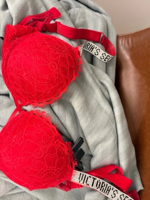 Röd spets-bh från Victoria's Secret - Snygg röd bh med vadderade kupor och spetsdetaljer från Victoria's Secret. Axelbanden är dekorerade med glittrande stenar och texten 'VICTORIA'S SECRET' för en extra lyxig känsla. Perfekt för dig som vill ha något extra. Några stenar fattas, se bild, annars bra kvalitet! 