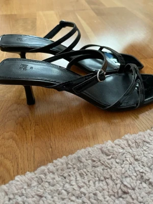 Svarta pumps med smala remmar från H&M - Snygga svarta pumps från H&M i lackmaterial med smala remmar och fyrkantig tå. Klackarna är smala och eleganta, och skorna har ett spänne vid ankeln. Perfekta för sommaren eller till en elegant utgång. Storlek 39. Skorna är använda 1 gång