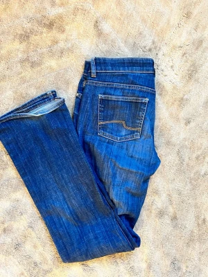 Blå bootcut jeans med fickdetalj - Snygga blå bootcut jeans med klassisk femficksdesign och kontrastsömmar på bakfickan. Jeansen har en något utsvängd passform och är tillverkade i denim med lätt tvättad look. Perfekta för en avslappnad och trendig stil.