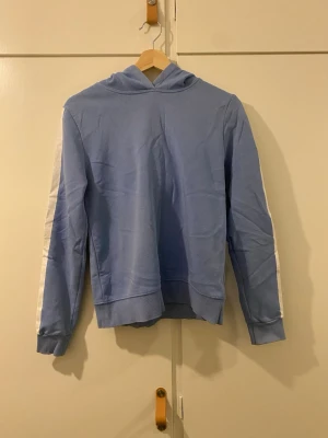Blå hoodie - Säljer denna hoodie med tryck på ärmarna. Fint skick utöver att trycket är lite ”urtvättat” men ger ett fint helhetsintryck! Något liten i storlek så är hellre storlek XS.