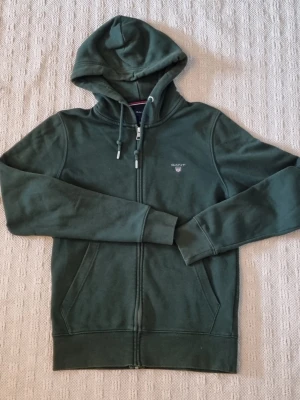 Grön hoodie med dragkedja från GANT - Mörkgrön hoodie från GANT med dragkedja framtill och justerbar huva med snören. Broderad GANT-logga och sköld på bröstet. Mjuk bomullsblandning och ribbade muddar vid ärmslut och nederkant. Två fickor framtill för extra chill känsla. Storlek small 