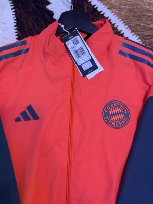 Röd FC Bayern München träningsjacka - Snygg röd träningsjacka från Adidas med FC Bayern München-logga på bröstet. Jackan har marinblå ärmar och detaljer samt dragkedja framtill. Klassiska Adidas-ränder på axlarna och tillverkad i lätt, sportigt material.