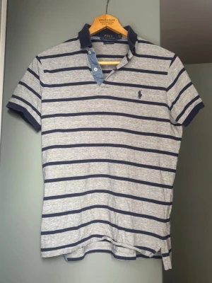 Randig pikétröja från Polo Ralph Lauren - Grå pikétröja med mörkblå horisontella ränder från Polo Ralph Lauren. Tröjan har klassisk krage, korta ärmar och knappslå med blå detalj. Broderad logga på bröstet och slim fit passform. Perfekt för en avslappnad men stilren look.