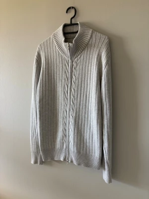 Ljusgrå ribbstickad kofta med dragkedja - Ljusgrå ribbstickad kofta med dragkedja framtill och hög krage. Koftan har ett diskret flätmönster längs dragkedjan och breda muddar vid ärmslut och nederkant. Perfekt för lager på lager och en clean look. Inget märke på den. Storlek M. Använd endast en gång