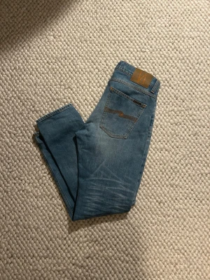 Ljusblå jeans från Nudie Jeans - Säljer ett par klassiska ljusblå jeans från Nudie Jeans med fem fickor och snygg design. Jeansen är slim fit i och är gjorda i mjukt denimtyg. Perfekta för dig som gillar en avslappnad och tidlös stil.