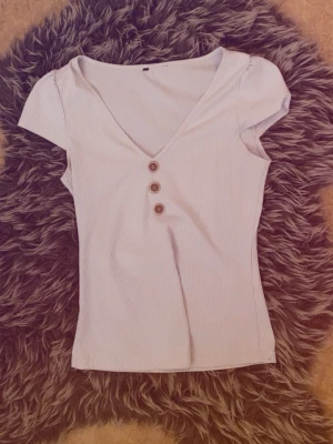 Vit ribbad topp med knappar - Snygg vit ribbad topp med v-ringning och tre dekorativa knappar framtill. Kortärmad och figurnära passform, perfekt att matcha med jeans eller kjol. Enkel men trendig design som passar till många olika stilar.