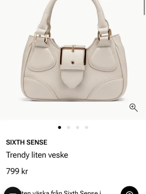 Beige handväska med guldspänne - Stilren beige handväska från Sixth Sense med unikt guldspänne framtill och dubbla handtag. Väskan har en mjuk, rundad form och är tillverkad i slätt konstläder. Snygga detaljer och modern design som lyfter din outfit.