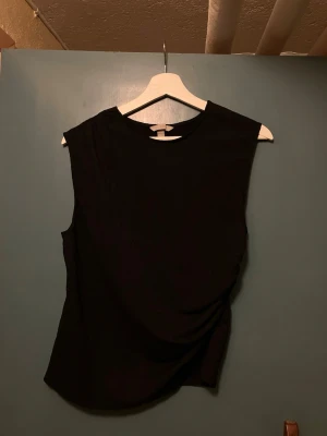 Svart ärmlös topp med drapering - Snygg svart ärmlös topp med rund halsringning och draperad detalj på sidan. Perfekt att styla med jeans eller kjol för en trendig look. Materialet känns mjukt och följsamt mot kroppen.