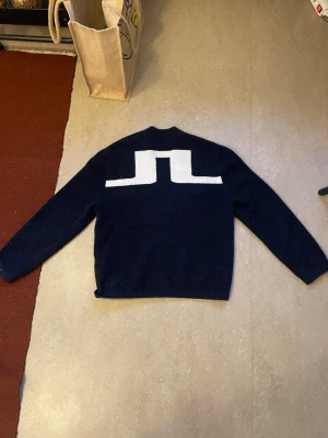 Mörkblå kofta från J.Lindeberg - Mysig mörkblå kofta från J.Lindeberg med stor vit logga på ryggen och broderade initialer 'S.L' framtill. Koftan har v-ringning, knappstängning och en bröstficka. Perfekt för lager på lager och stilren look.