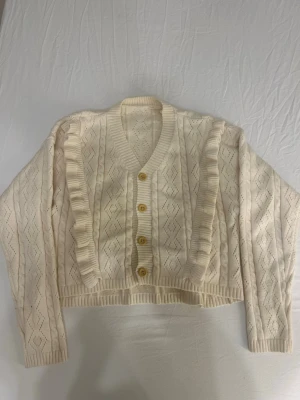 Krämvit stickad kofta med volang - Söt krämvit stickad kofta med dekorativt hålmönster och volangdetaljer längs framkanterna. Koftan har v-ringning, ribbade muddar och fyra beige knappar framtill. Kortare modell som ger en trendig look. Materialet ser ut att vara mjukt och bekvämt, troligen bomull eller akryl.