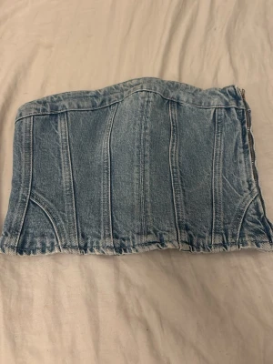 Ljusblå denim korsett tubtopp - Trendig ljusblå denim korsett tubtopp med markerade sömmar och dragkedja i sidan. Snygg att styla med både jeans och kjol för en cool look. Klassisk jeansdetalj och croppad passform.
