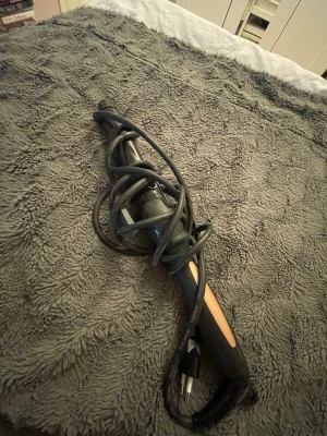 Svart locktång med klämma - En svart locktång med klämma och sladd, perfekt för att skapa snygga lockar. Handtaget har en beige detalj och tången är tillverkad i plast och metall. Passar dig som vill styla håret enkelt hemma.