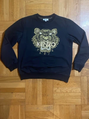 Svart sweatshirt från Kenzo med tiger - Säljer en svart sweatshirt från Kenzo med det ikoniska tigerhuvudet broderat i guld och vitt på bröstet samt texten 'KENZO PARIS'. Tröjan har rund hals, långa ärmar och ribbade muddar. Perfekt för dig som vill sticka ut med en klassisk streetwear-look.