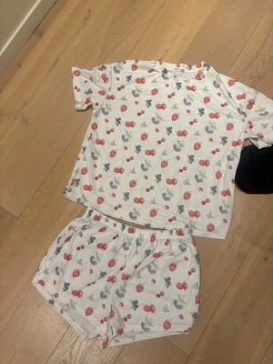 Vit pyjamas med bärmönster - Söt tvådelad pyjamas i vitt med kortärmad t-shirt och shorts. Setet har ett färgglatt bärmönster med jordgubbar, blåbär och hallon i rött, lila och grönt. Mjuk och skön bomullskänsla, perfekt för mysiga kvällar hemma. Den är i strlk L men skulle säga ett den är i en medium eller S istället!